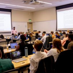 Students sit in a lecture hall with their laptops while their instructor points to the lecture on a screen. 