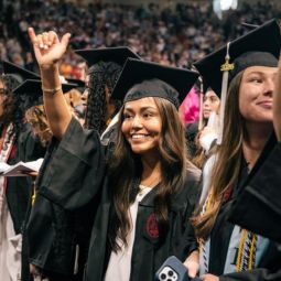A female student wearing her black cap and gown is smiling while doing the spurs up hand gesture. 