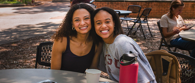 Two students hangout at a table outside of the Russell House, the Thomas Cooper fountain can be seen in the background.