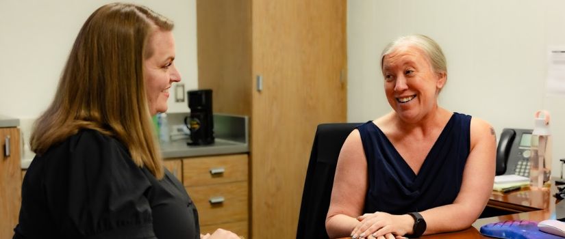 two employees sitting at a desk talking to one another 