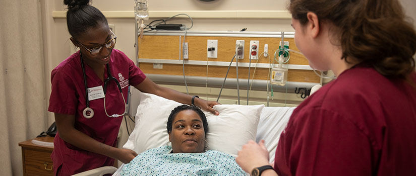 A simulation hospital room with two nursing students attending a female actor in the role of a patient in a hospital bed